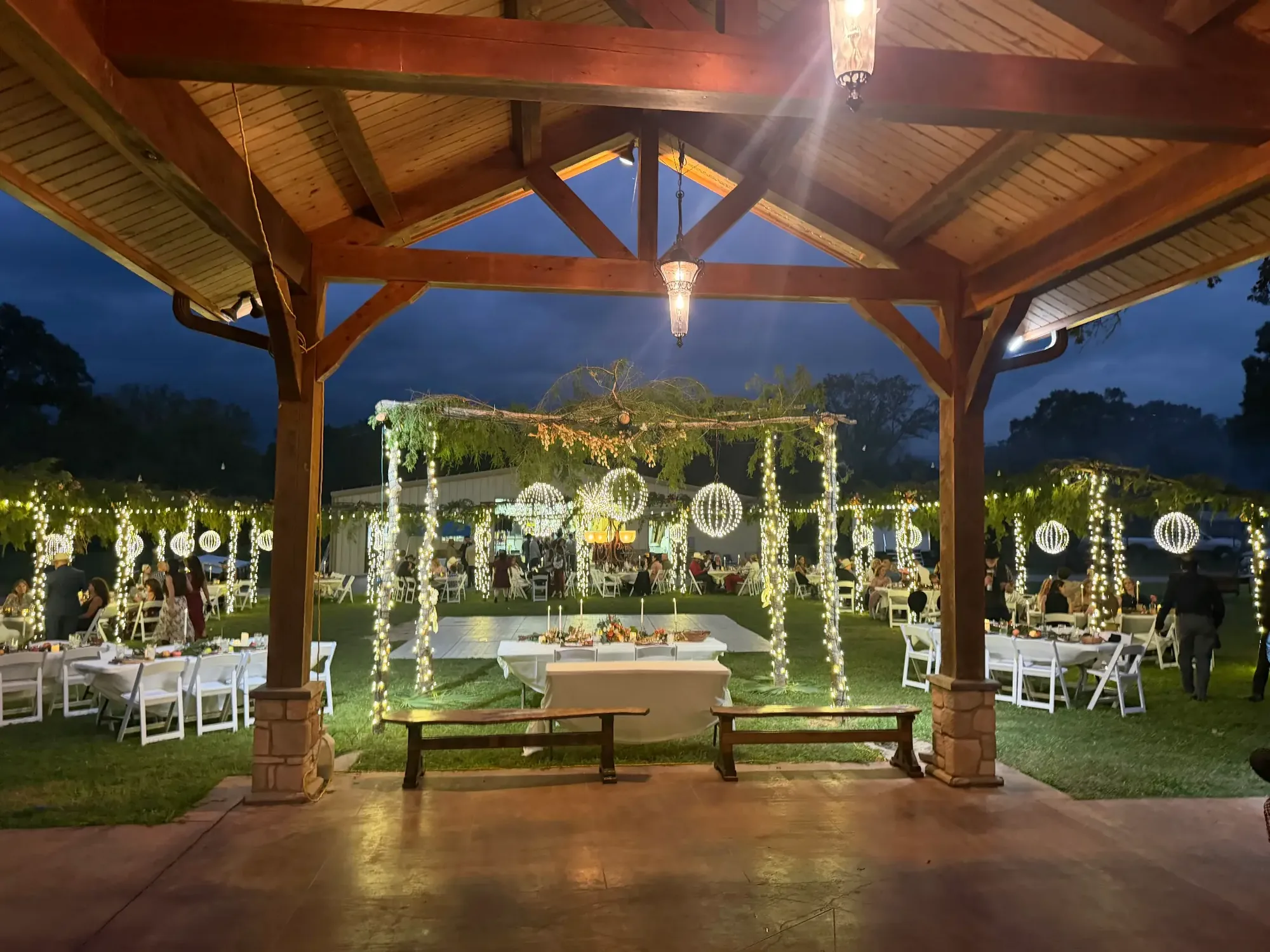 Timber pavilion at night with a lit evergreen garland arch, wrapped columns and banqueting tables set beneath it.