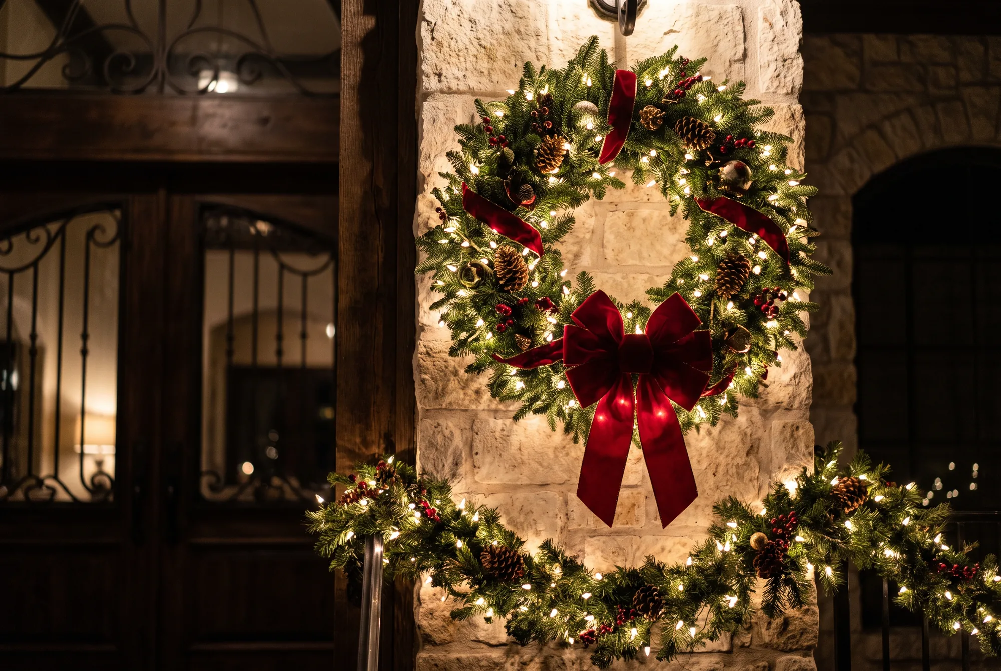 Large illuminated evergreen wreath with a red velvet bow mounted on a Texas limestone column beside lit garland.