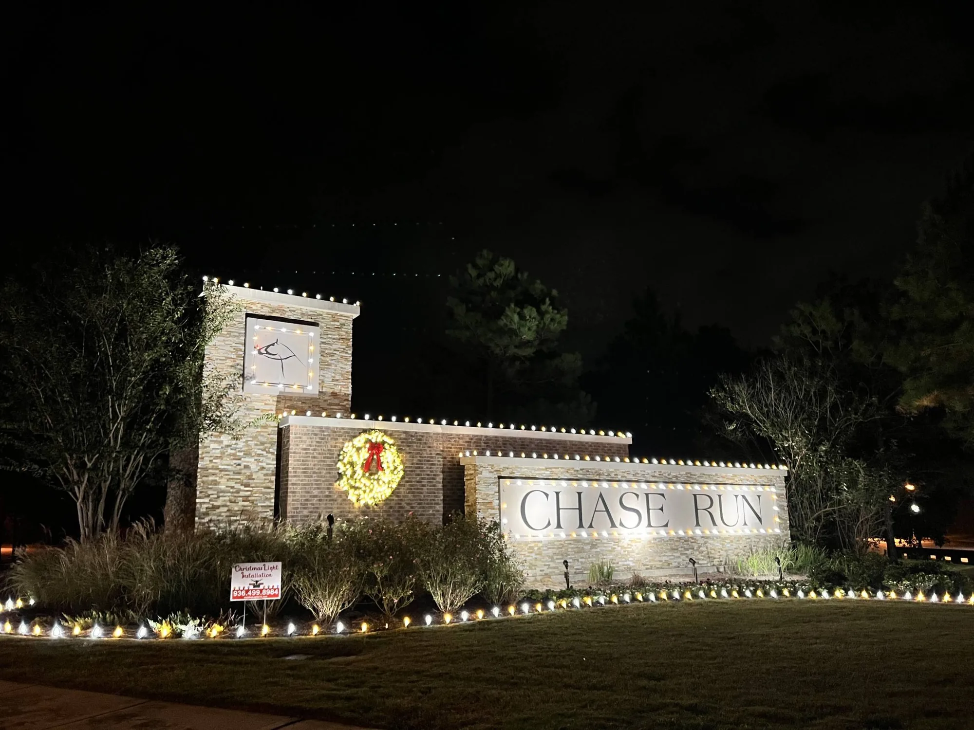 Community monument entrance at night with a lit wreath, a lit stone tower and warm-white planting-bed lighting.