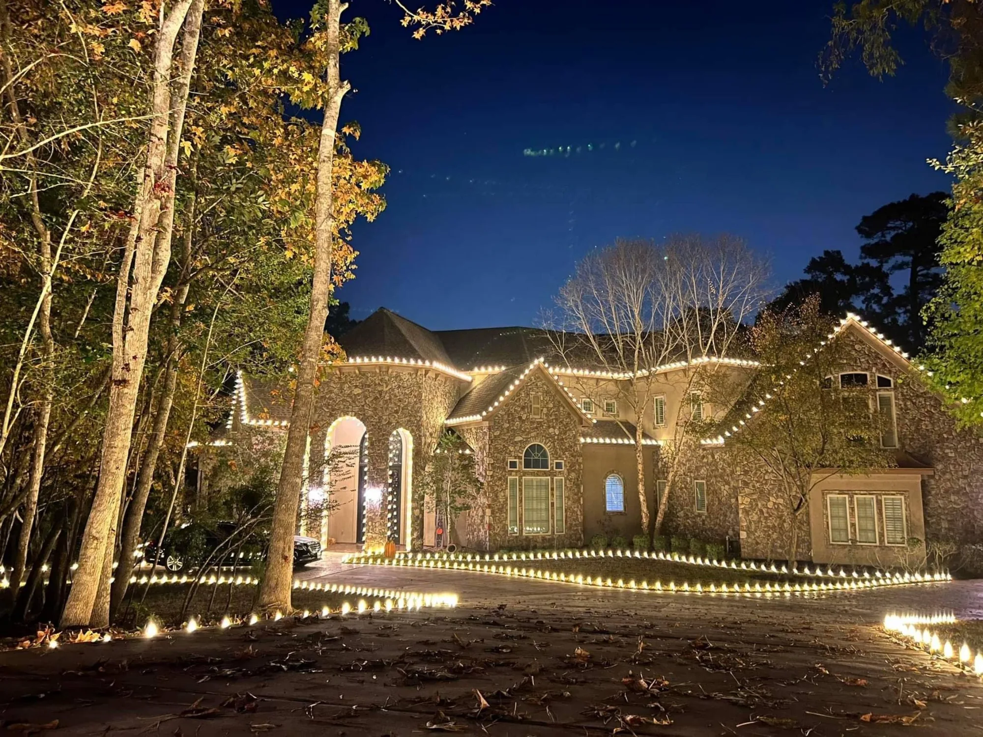 Large stone Texas estate at night with a lit driveway edge, wrapped tree trunks and warm-white rooflines.